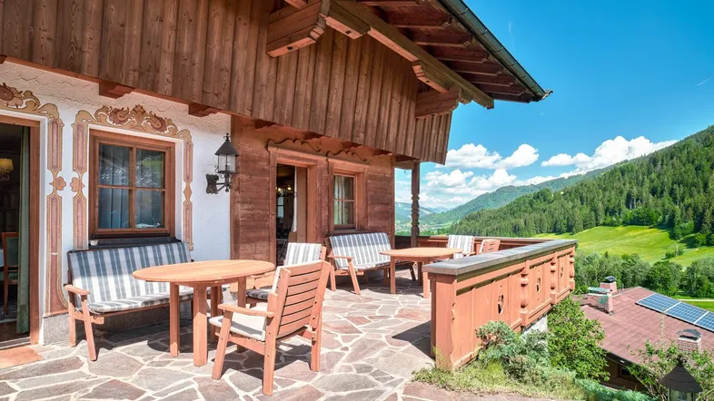 Große Steinterrasse mit Holzmöbeln und traditioneller Fassadenmalerei, umgeben von grünen Bergen.