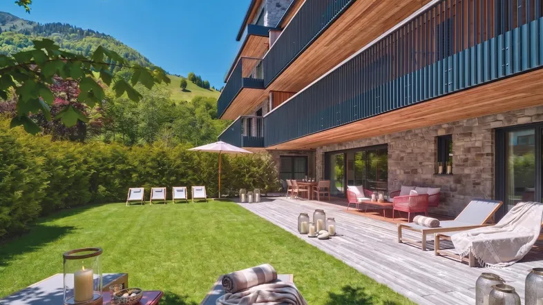 Moderne Wohnanlage mit Holzfassade, großer Rasenfläche und möblierter Holzterrasse mit Bergblick.