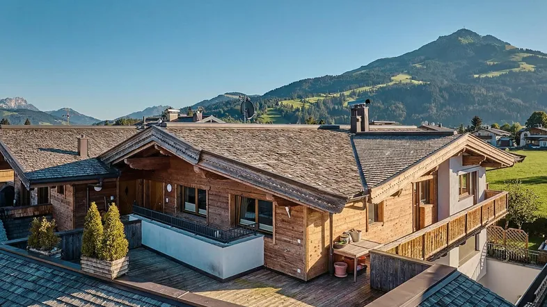 Luftaufnahme eines traditionellen Holzchalets mit mehreren Balkonen und Dächern vor einer malerischen Bergkulisse.