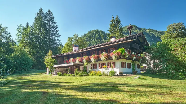 Traditionelles Alpenhaus mit Blumenkästen, umgeben von einer weitläufigen Grünfläche und Bergen im Hintergrund.