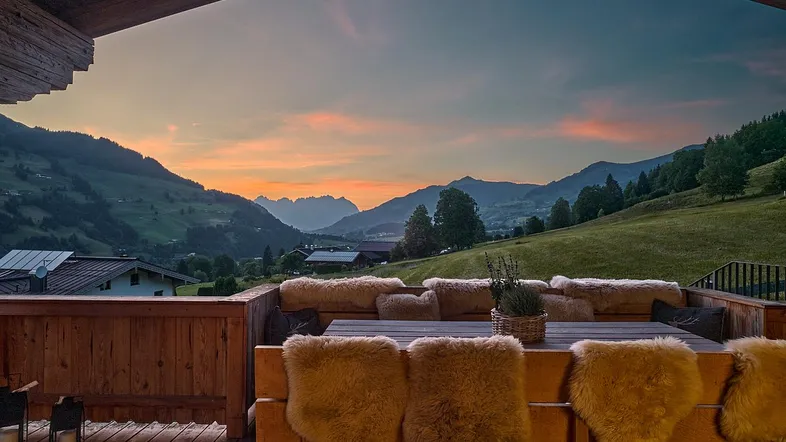 Gemütliche überdachte Terrasse mit Holzbänken, Fellen und atemberaubendem Bergblick bei Sonnenuntergang.