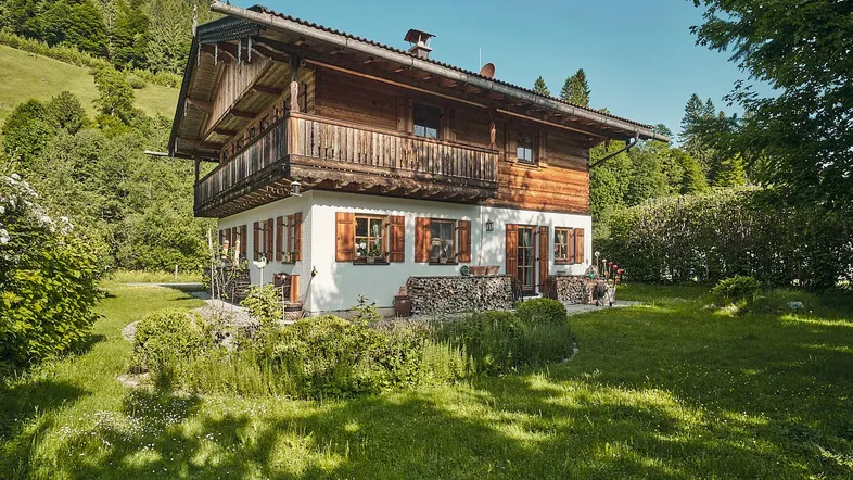Traditionelles Holzhaus im Chalet-Stil mit weißer Fassade, umgeben von grüner Natur und Bergen.