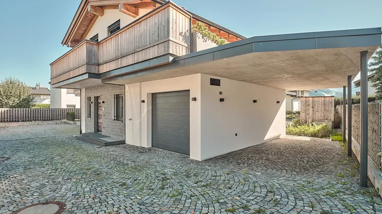 Modernes Haus mit Carport, Garage und gepflasterter Auffahrt, umgeben von grüner Landschaft.