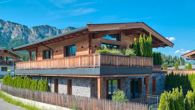 Modernes Chalet mit Holzfassade, Steinmauerwerk und großem Balkon vor alpiner Bergkulisse.