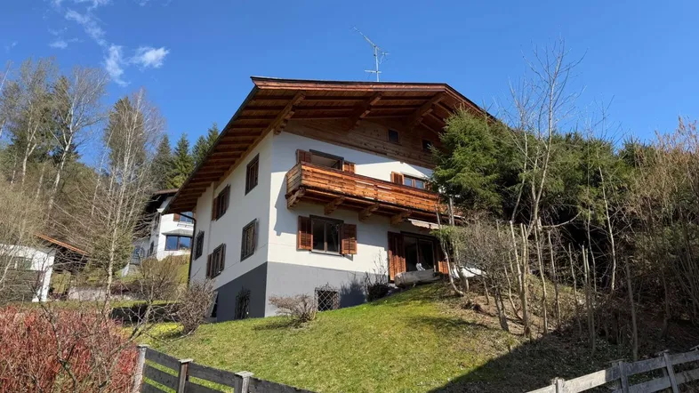 Traditionelles Haus am Hang mit Holzbalkon und grüner Umgebung unter blauem Himmel.