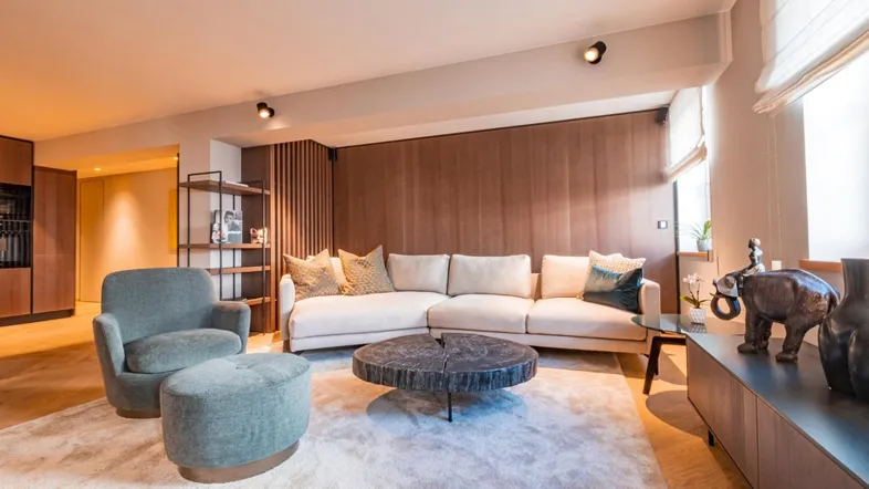 Modernes Wohnzimmer mit beigem Sofa, Holzakzenten und stilvollem Couchtisch auf hellem Teppich.