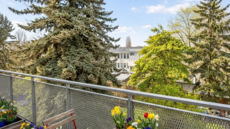 Gemütlicher Balkon mit Holztisch, Stühlen und bunten Blumen, umgeben von hohen Bäumen und viel Grün.