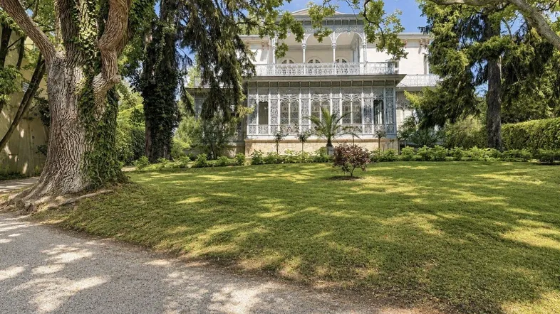 Prächtiges historisches Gebäude mit weitläufigem Garten und altem Baumbestand unter blauem Himmel.