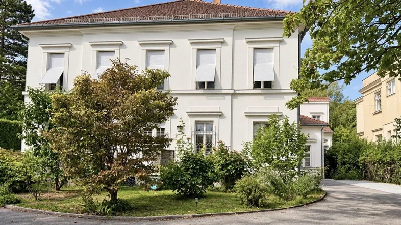 Großes weißes Gebäude mit klassischer Architektur, umgeben von Bäumen und einem gepflegten Vorgarten.