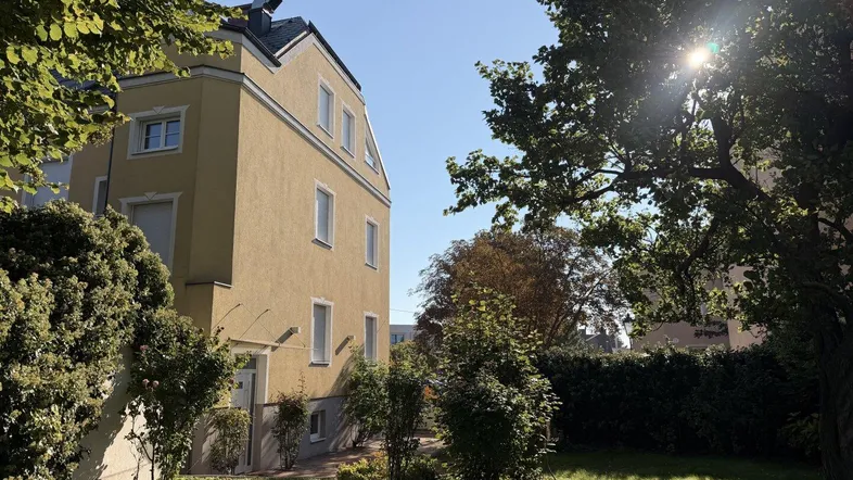 Gelbes Mehrfamilienhaus mit grüner Gartenanlage und Bäumen unter strahlendem Sonnenschein.