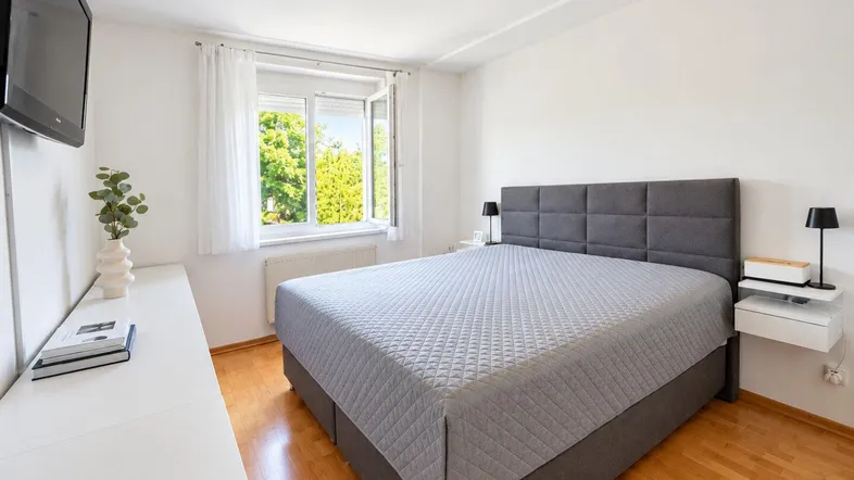 Helles Schlafzimmer mit Holzboden, großem Bett und Blick ins Grüne durch das Fenster.