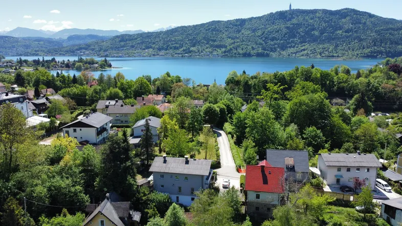 Panoramablick auf eine Wohnsiedlung am Seeufer mit bewaldeten Hügeln und Bergen im Hintergrund.