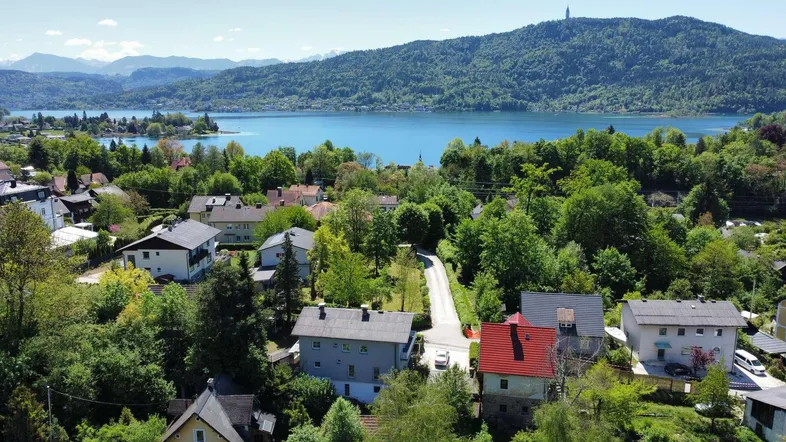 Luftaufnahme einer Wohnsiedlung mit Häusern, umgeben von viel Grün, einem See und Bergen im Hintergrund.