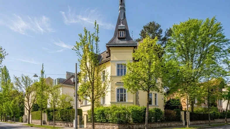 Historische Villa mit markantem Turm und gepflegter Gartenanlage an einer Straßenecke.