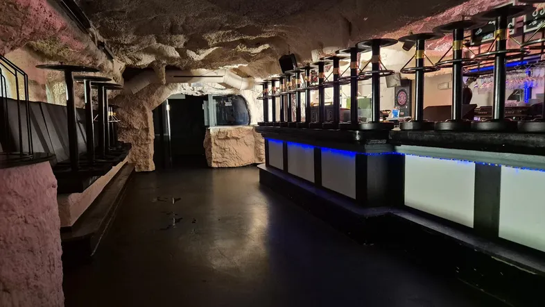 Dunkler Barbereich mit Höhlen-Design, hohen Tischen und blauer LED-Beleuchtung, ideal für Veranstaltungen.