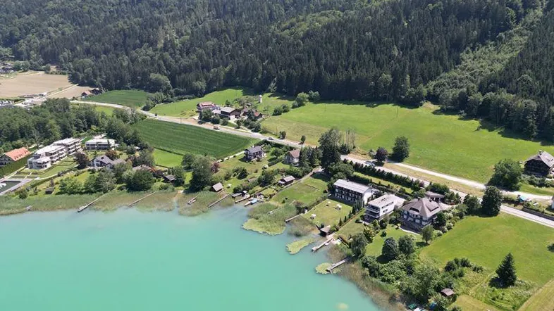 Luftaufnahme eines malerischen Sees mit Häusern am Ufer und bewaldeten Bergen im Hintergrund.