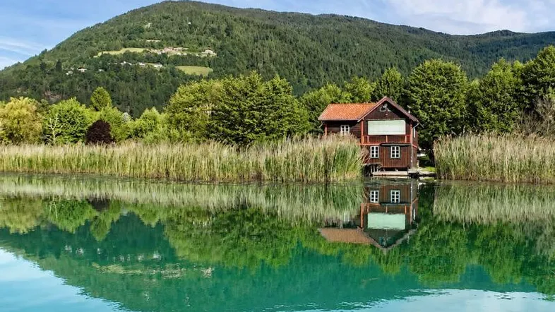 Idyllisches Haus am Seeufer mit Schilf und bewaldeten Bergen im Hintergrund, spiegelt sich im klaren Wasser.