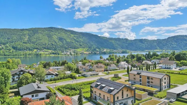 Panoramablick auf moderne Neubauten mit Photovoltaikanlagen, umgeben von grüner Landschaft und einem See mit Bergen im Hintergrund.