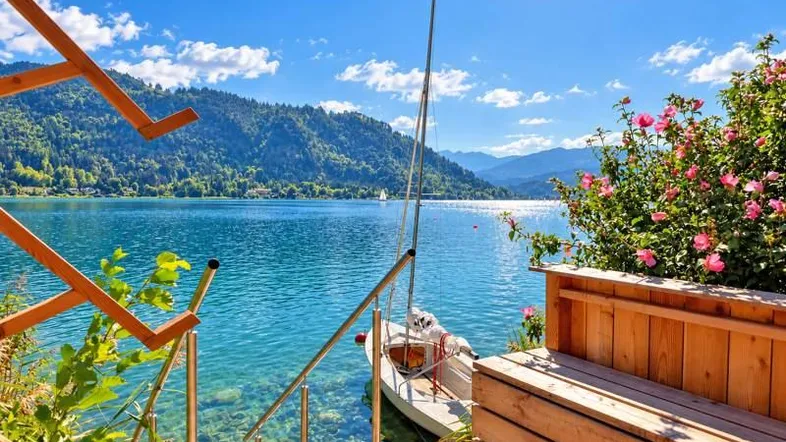 Idyllisches Seeufer mit Holzsteg, Treppe zum Wasser und Segelboot vor malerischer Bergkulisse.
