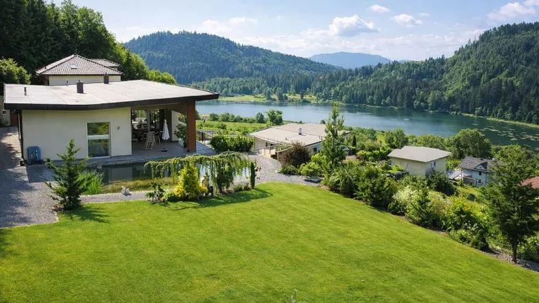 Panoramablick auf das moderne Haus mit großem Garten, Naturpool und See im Hintergrund.