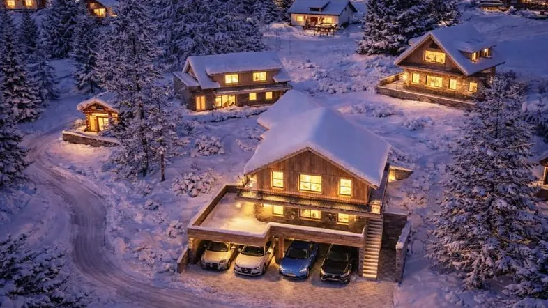 Winterliche Ansicht von mehreren Chalets in einer verschneiten Berglandschaft bei Abenddämmerung mit beleuchteten Fenstern und Garagen.