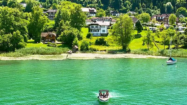 Panoramablick auf den See mit Häusern am Ufer und bewaldeten Hügeln im Hintergrund.
