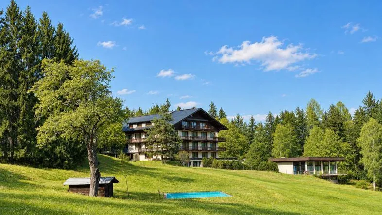 Großes Wohngebäude auf einem grünen Hügel mit einem kleinen Pool und umgeben von Bäumen unter blauem Himmel.