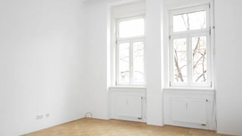 Heller, leerer Raum mit zwei großen Fenstern und klassischem Parkettboden.