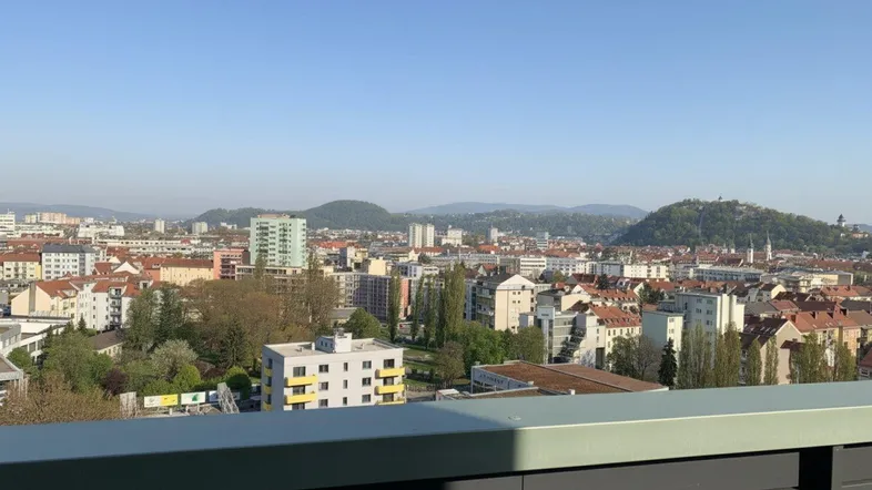 Panoramablick über die Stadt mit Gebäuden und grünen Hügeln im Hintergrund unter blauem Himmel.
