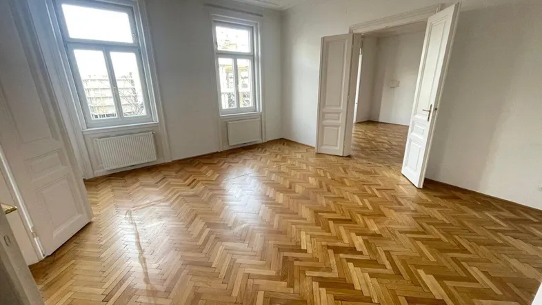 Geräumiges Zimmer mit Fischgrätparkett, hohen Decken und großen Fenstern, das viel Tageslicht hereinlässt.