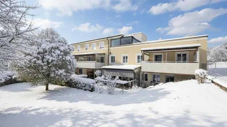 Gebäudeansicht im Winter mit schneebedecktem Garten und Balkonen unter blauem Himmel.