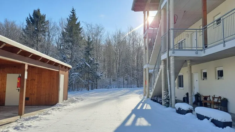 Gebäudeansicht im Winter mit verschneiter Umgebung und Wald im Hintergrund.