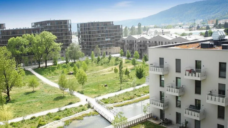 Luftaufnahme moderner Wohngebäude mit Grünflächen, einem Kanal und Bergen im Hintergrund.