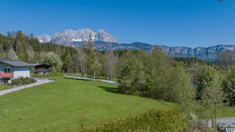 Weitläufige grüne Landschaft mit Häusern, Bäumen und majestätischen Bergen im Hintergrund unter blauem Himmel.