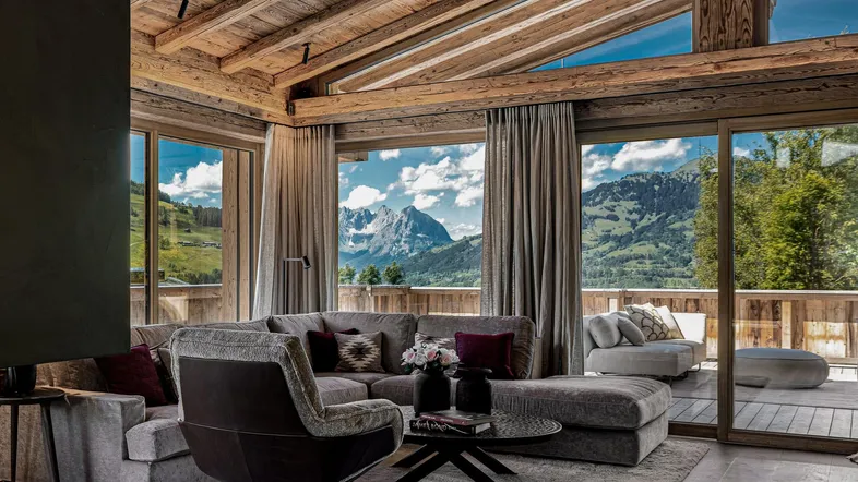 Geräumiges Wohnzimmer im Chalet-Stil mit Holzbalkendecke, großen Fenstern und atemberaubendem Bergblick.