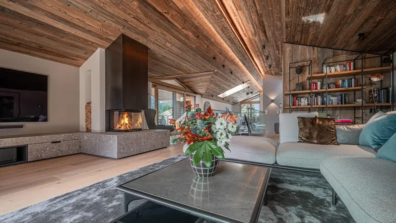 Gemütliches Wohnzimmer mit Holzbalkendecke, modernem Kamin und Blick ins Grüne durch große Fenster.