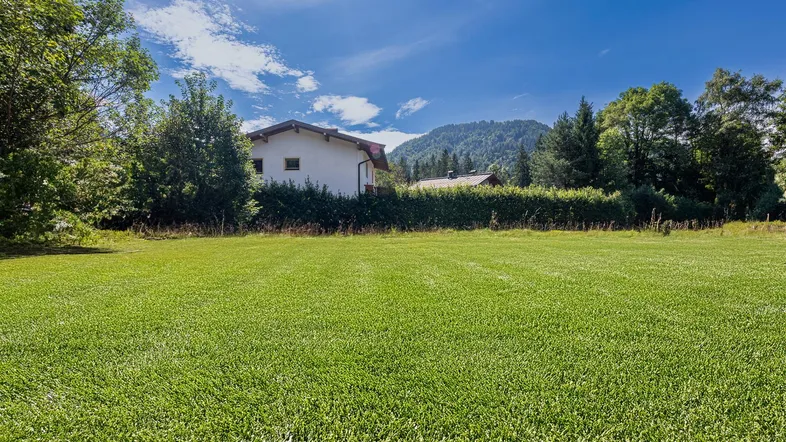 Weitläufige Grünfläche mit Blick auf ein Wohnhaus und die umliegende Berglandschaft unter blauem Himmel.