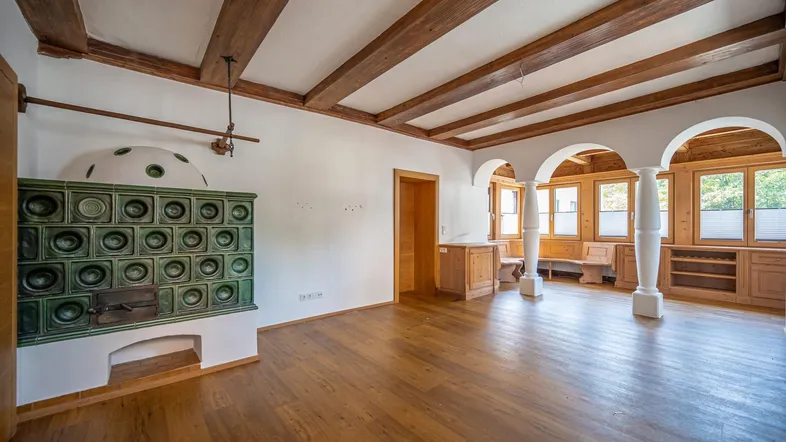 Geräumiges Wohnzimmer mit traditionellem Kachelofen, Holzbalkendecke und hellem Parkettboden.