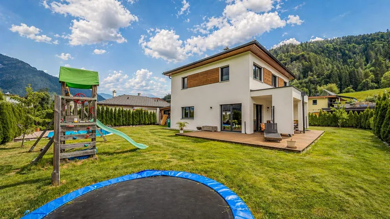 Modernes Einfamilienhaus mit großem Garten, Holzterrasse, Spielplatz und Trampolin vor Bergkulisse.