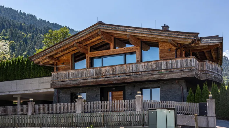 Modernes Holzhaus mit traditionellen Elementen, großen Fenstern und Balkonen vor bewaldeter Bergkulisse.