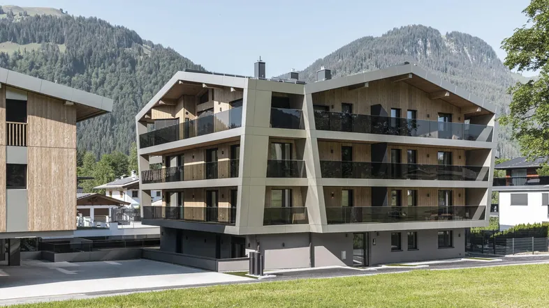 Modernes Apartmentgebäude mit Holzfassade und Balkonen vor alpiner Bergkulisse.