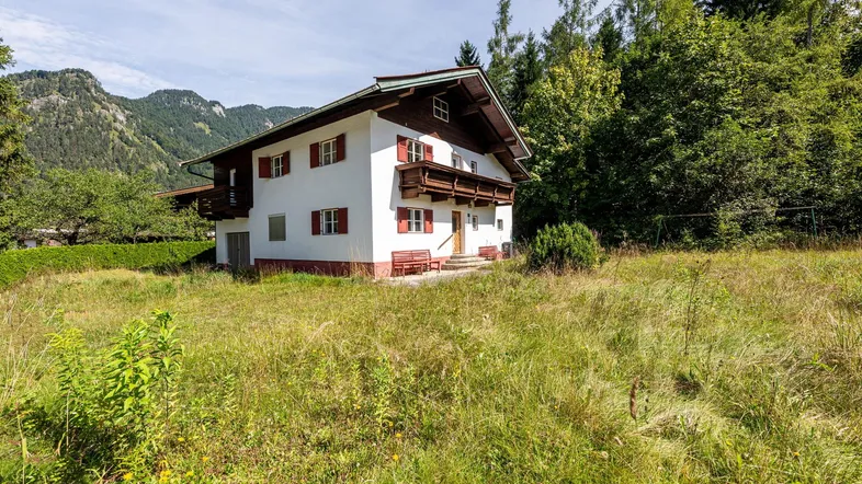 Traditionelles Haus im alpenländischen Stil mit roten Fensterläden und Holzbalkonen, umgeben von Natur und Bergen.