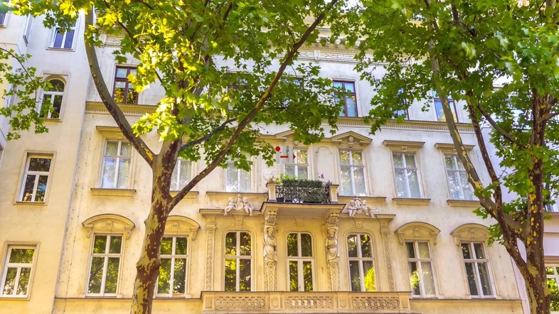 Historische Altbaufassade mit Stuckverzierungen, kleinem Balkon und grünen Bäumen davor.