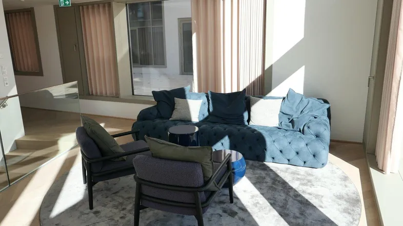 Gemütlicher Loungebereich mit bequemen blauen Sofas, Sesseln und einem runden Teppich, hell durch große Fenster.