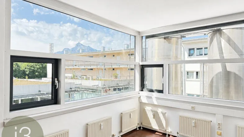 Heller Raum mit großen Fenstern, dunklem Holzboden und weitem Blick auf Berge und umliegende Gebäude.
