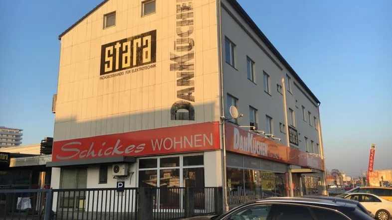 Gebäudeansicht eines Geschäftshauses mit Werbeschildern und geparkten Autos davor, unter klarem Himmel.