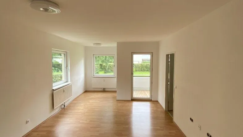 Helles, leeres Wohnzimmer mit Laminatboden und Zugang zu einem Balkon, bietet viel natürliches Licht.