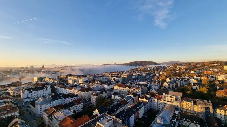 Panoramablick über eine Stadtlandschaft mit Nebel in den Tälern und Bergen im Hintergrund.