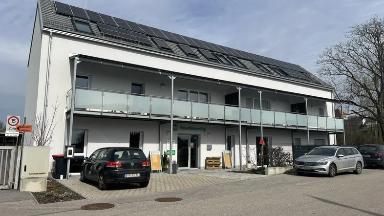 Modernes Geschäftsgebäude mit Solarpaneelen auf dem Dach und Parkplätzen davor, umgeben von Bäumen.