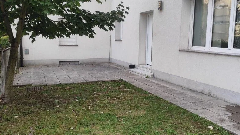 Seitenansicht des Gebäudes mit Eingangsbereich, gepflastertem Weg und kleiner Grünfläche.